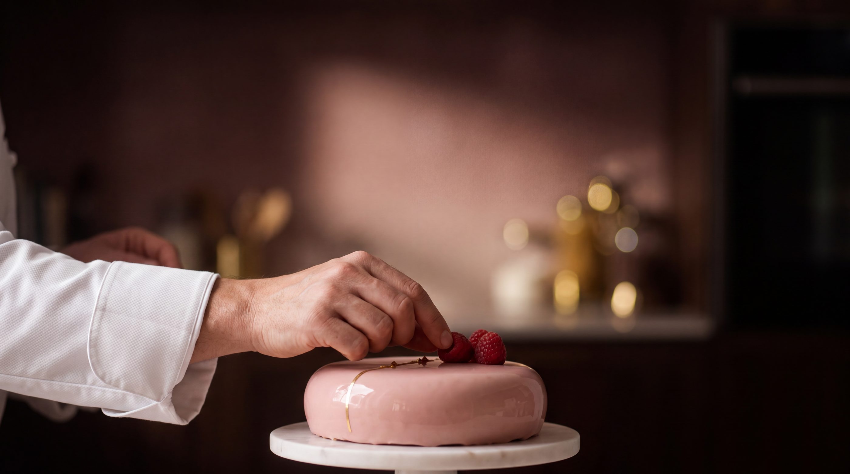 A pâtissier places a raspberry on a pink mirror-glazed entremet